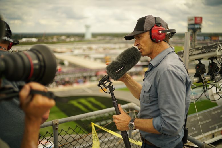 Concord, NC - Host Phil Keoghan (R) speaks into a microphone while filming above the Charlotte Motor Speedway's Roval course. (National Geographic/Alex Mandiola)