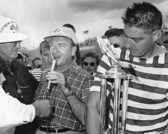 DAYTONA BEACH, FL — February 12, 1960: Walter Cronkite of CBS Television (C) gets set to interview Fireball Roberts (R) after Roberts won the first qualifying race for the upcoming Daytona 500 NASCAR Cup race at Daytona International Speedway. NASCAR Commissioner Erwin “Cannon Ball” Baker (L) looks on. (Photo by ISC Images & Archives via Getty Images)