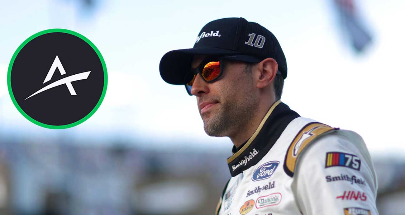 Aric Almirola looks on in Smithfield fire suit and hat with The Action Network graphic in upper-left of photo