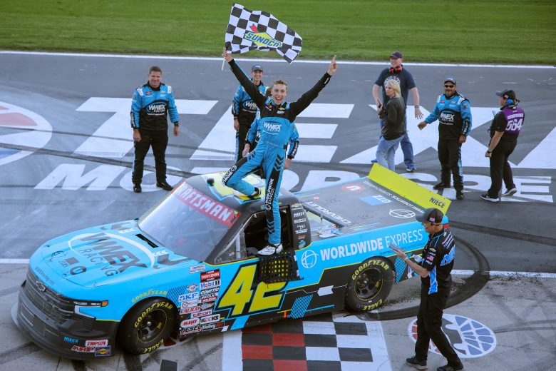 Carson Hocevar stands on his parked truck at Victory Lane, holding the checkered flag with his hands in the air