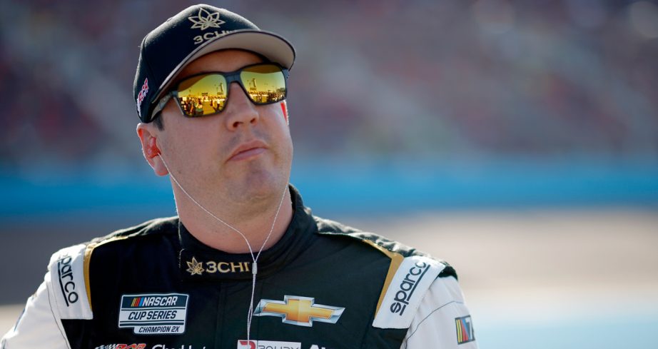 Kyle Busch stands on pit road looking into the distance at Phoenix Raceway wearing a black hat and orange-tinted sunglasses.
