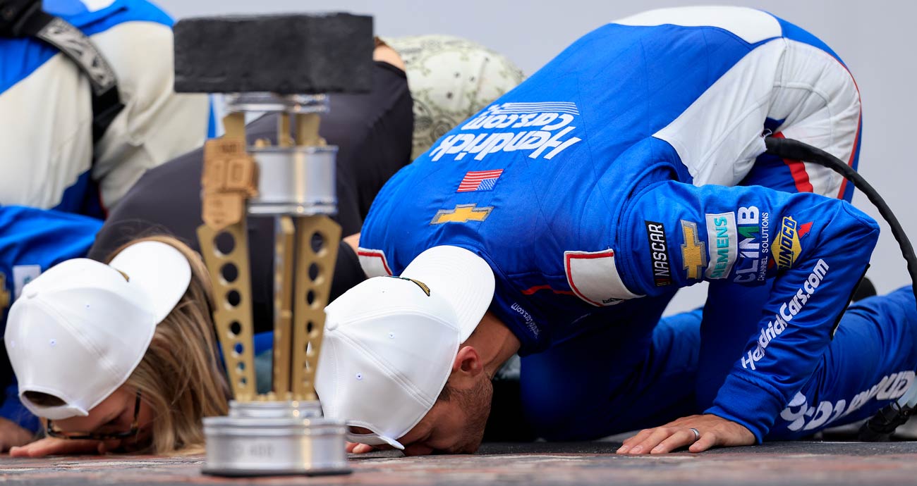 Kyle Larson kisses the Indianapolis Motor Speedway bricks as the winner's trophy sits in the foreground.