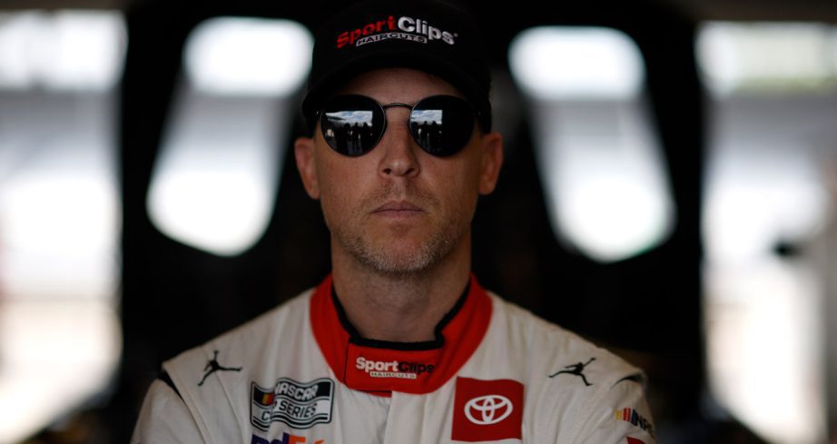 Denny Hamlin, in sunglasses, looks straight ahead in a dimly lit NASCAR garage.