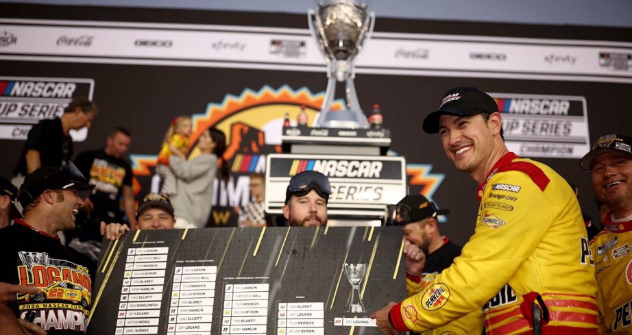 Joey Logano points to his name in the NASCAR Cup Series Playoffs Grid challenge bracket as the Bill France Cup sits in the background.