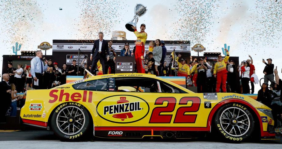 Joey Logano hoists the Bill France Cup in Victory Lane as his No. 22 Team Penske Ford is parked in the foreground at Phoenix Raceway.