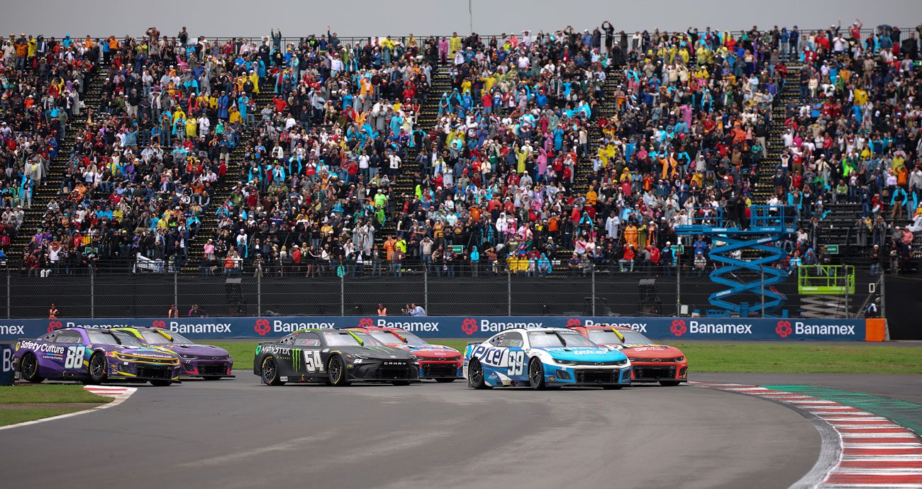 NASCAR Cup Series cars race through a turn at Autodromo Hermanos Rodriguez as the crowd watches in the background.