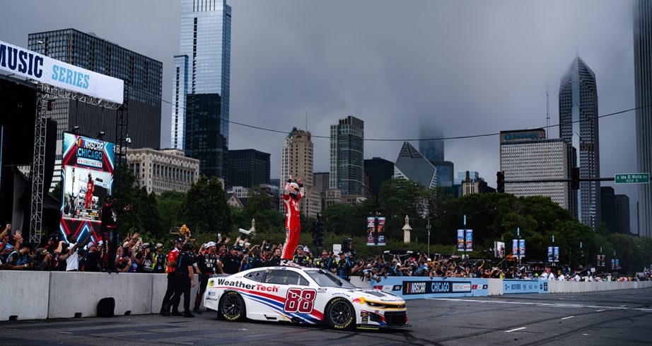 Shane van Gisbergen stands on top of No. 88 Weather Tech car and celebrates in front of crowd with Chicago Skyline in the background.