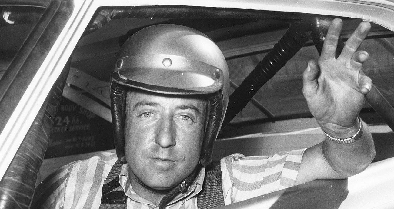 Rex White smiles and waves from the driver seat of his car with a helmet