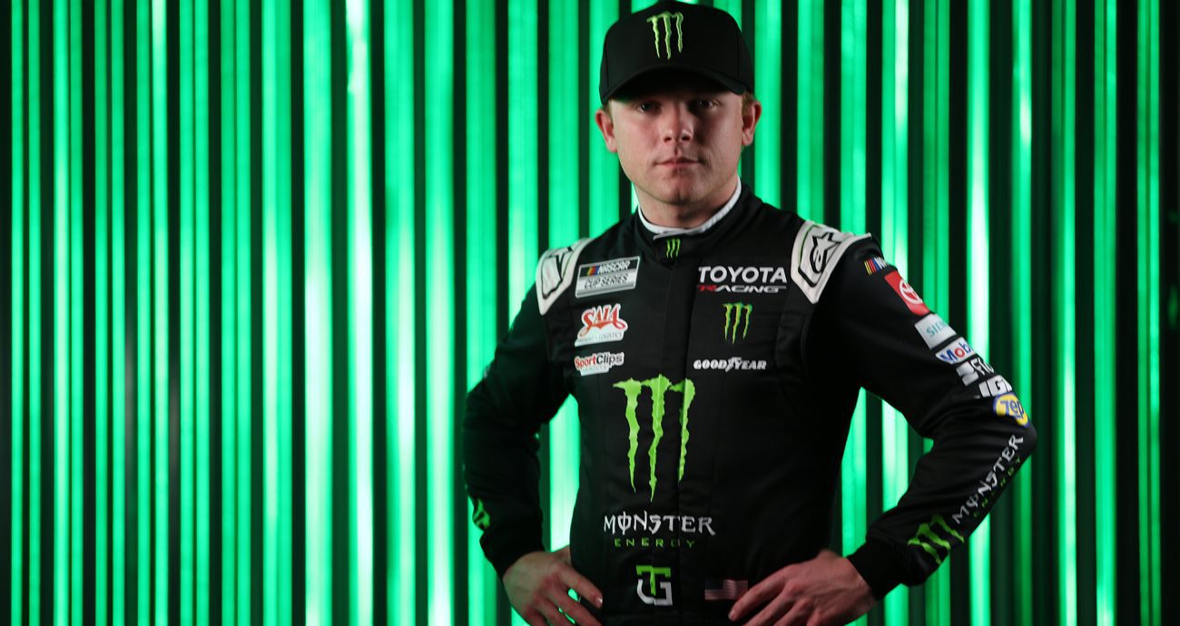 Ty Gibbs stands with hands on hips in front of a green background wearing his Monster Energy fire suit.