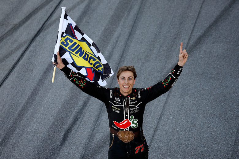 Carson Hocevar holds the checkered flag and points to the sky.