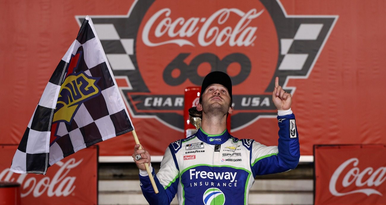 Daniel Suarez points up to the sky while holding the checkered flag in Victory Lane.