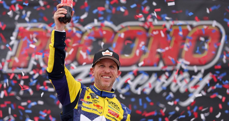 Denny Hamlin raises his right arm while holding a bottle of Coca-Cola as he celebrates in Victory Lane on June 14, 2026 at Pocono Raceway.
