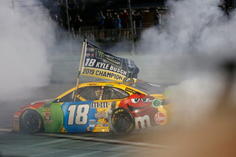 Kyle Busch does a celebratory burnout in his No. 18 Joe Gibbs Racing Toyota while holding a championship flag.